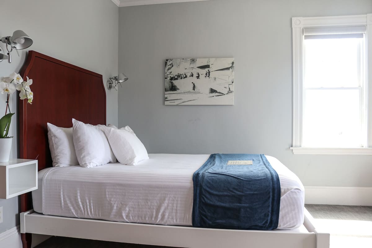 A bedroom with a bed, white walls, a wooden headboard, a framed abstract painting, and a window.