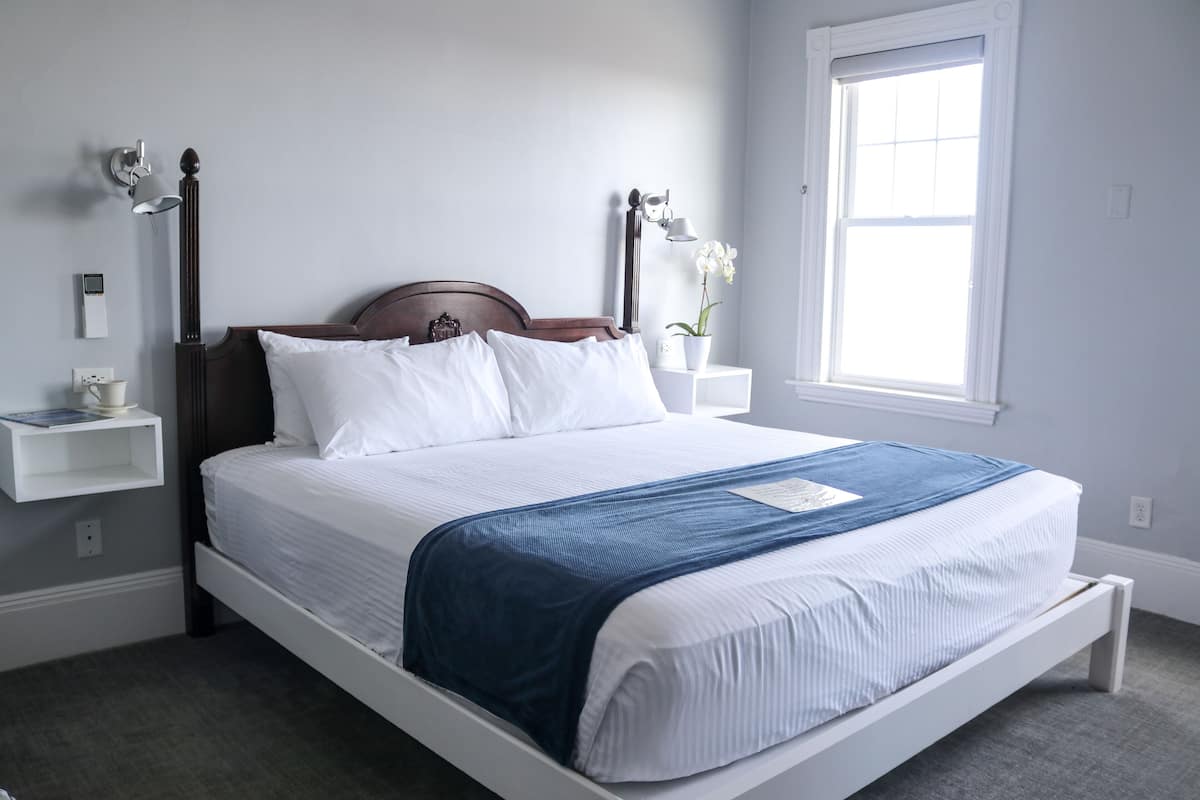 A neatly made bed with white and blue bedding, a wooden headboard, and a bedside table with a lamp and a small plant.