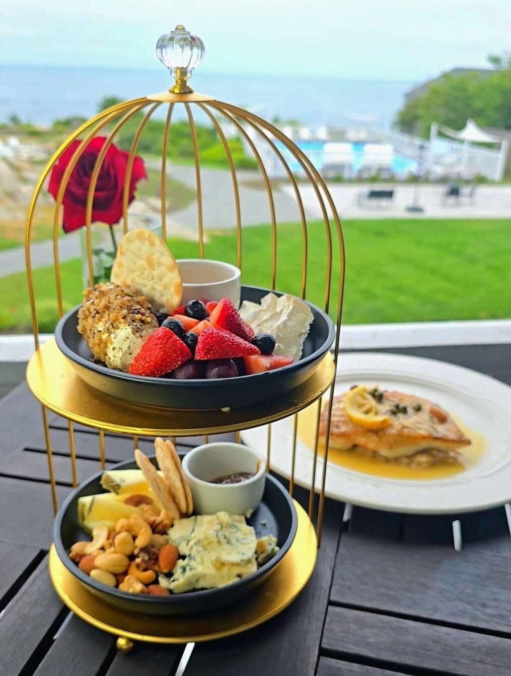 A three-tiered dessert tray with various sweets and fruits, a single rose, and a pastry on a table with a view of the sea.
