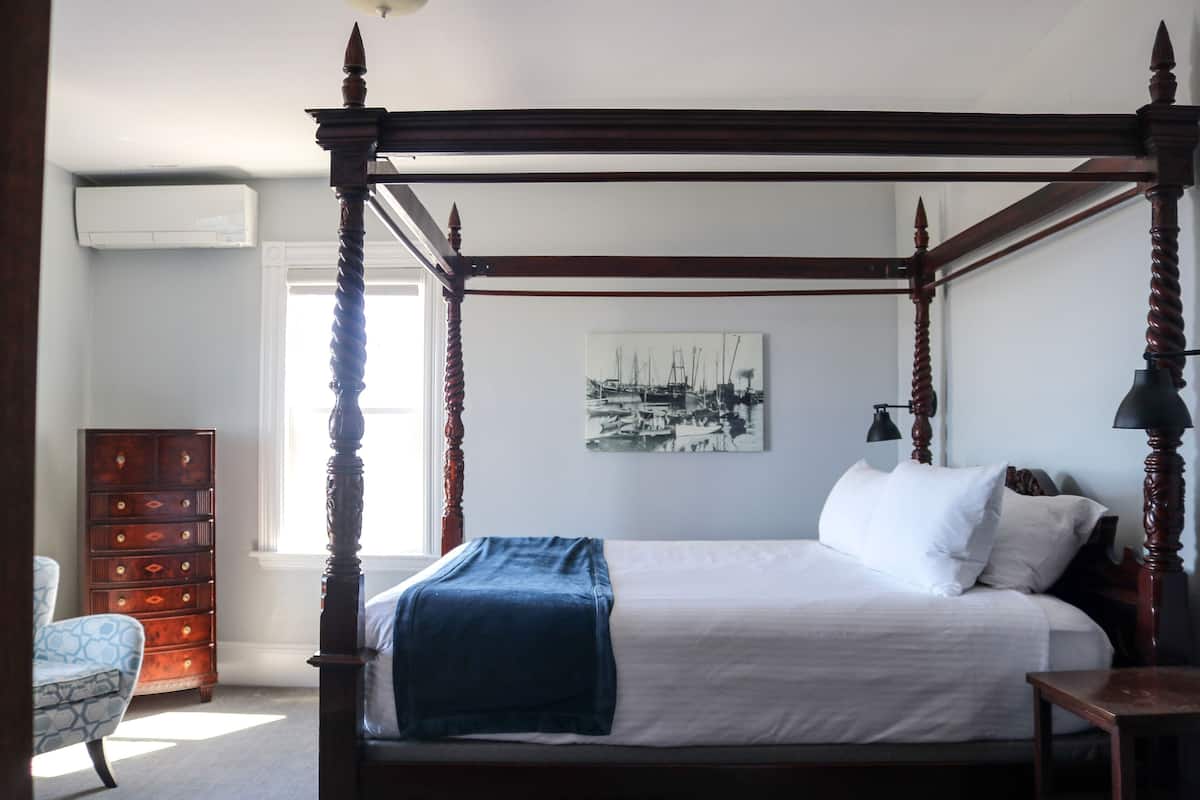 A four-poster bed with a blue throw blanket, a wooden dresser, and a chair in a room with a white wall and a black-and-white photograph.