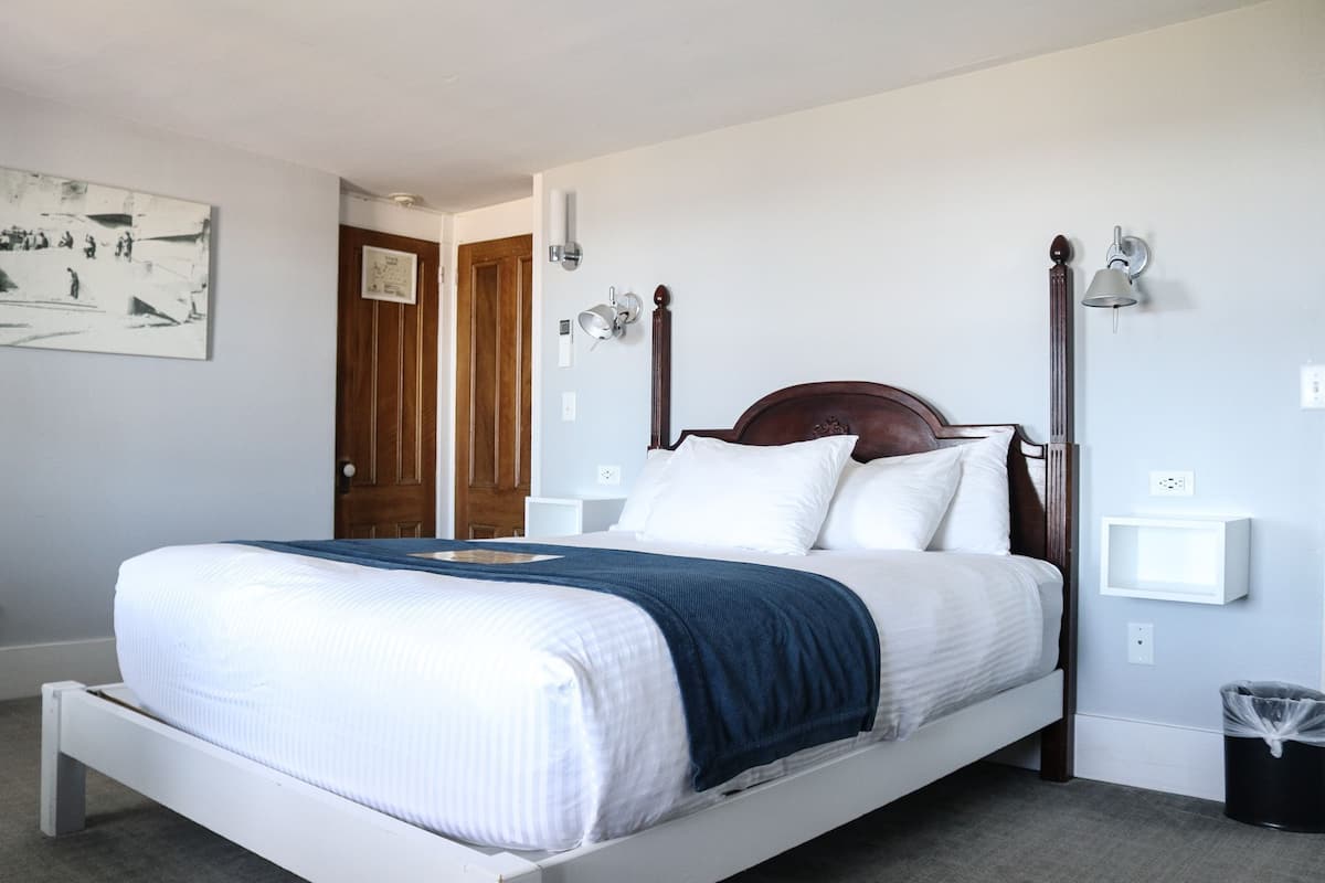 A neatly made bed with a wooden headboard, white bedding, and a blue throw blanket. There are two wall-mounted nightstands with lamps, a black trash bin, and a framed picture on the wall.