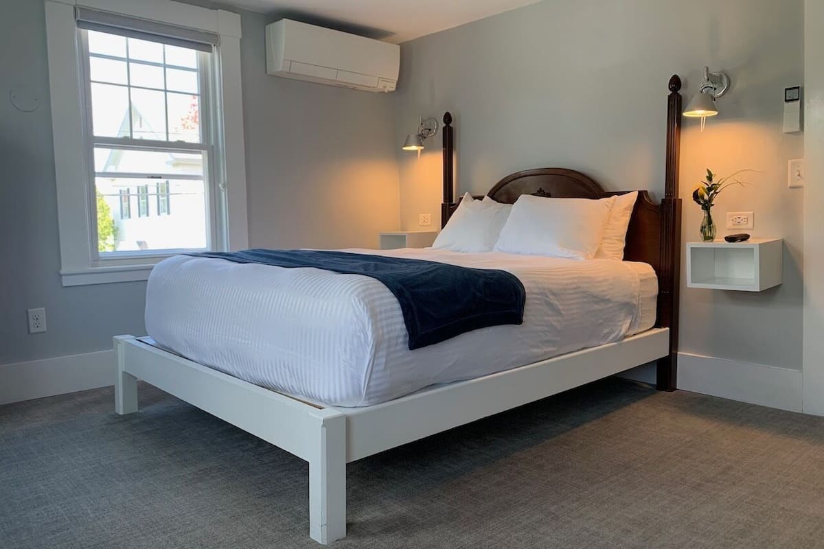 A neatly made bed with white linens and a dark throw blanket, a bedside table with a vase, and a window with blinds.