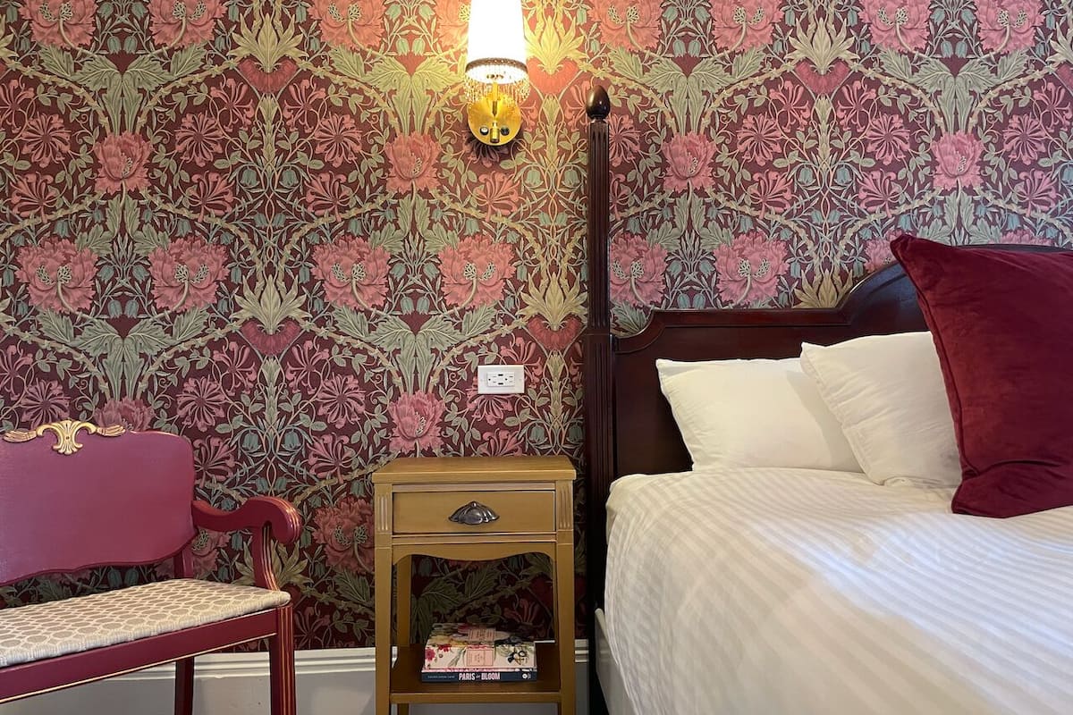 A bedroom with a bed, a nightstand, a chair, and a wall with floral wallpaper.