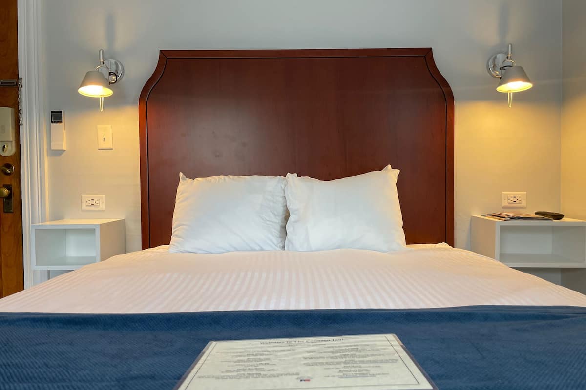 A neatly made bed with white linens and two pillows, flanked by two bedside tables with lamps, in a room with a wooden headboard and a wall-mounted light fixture.