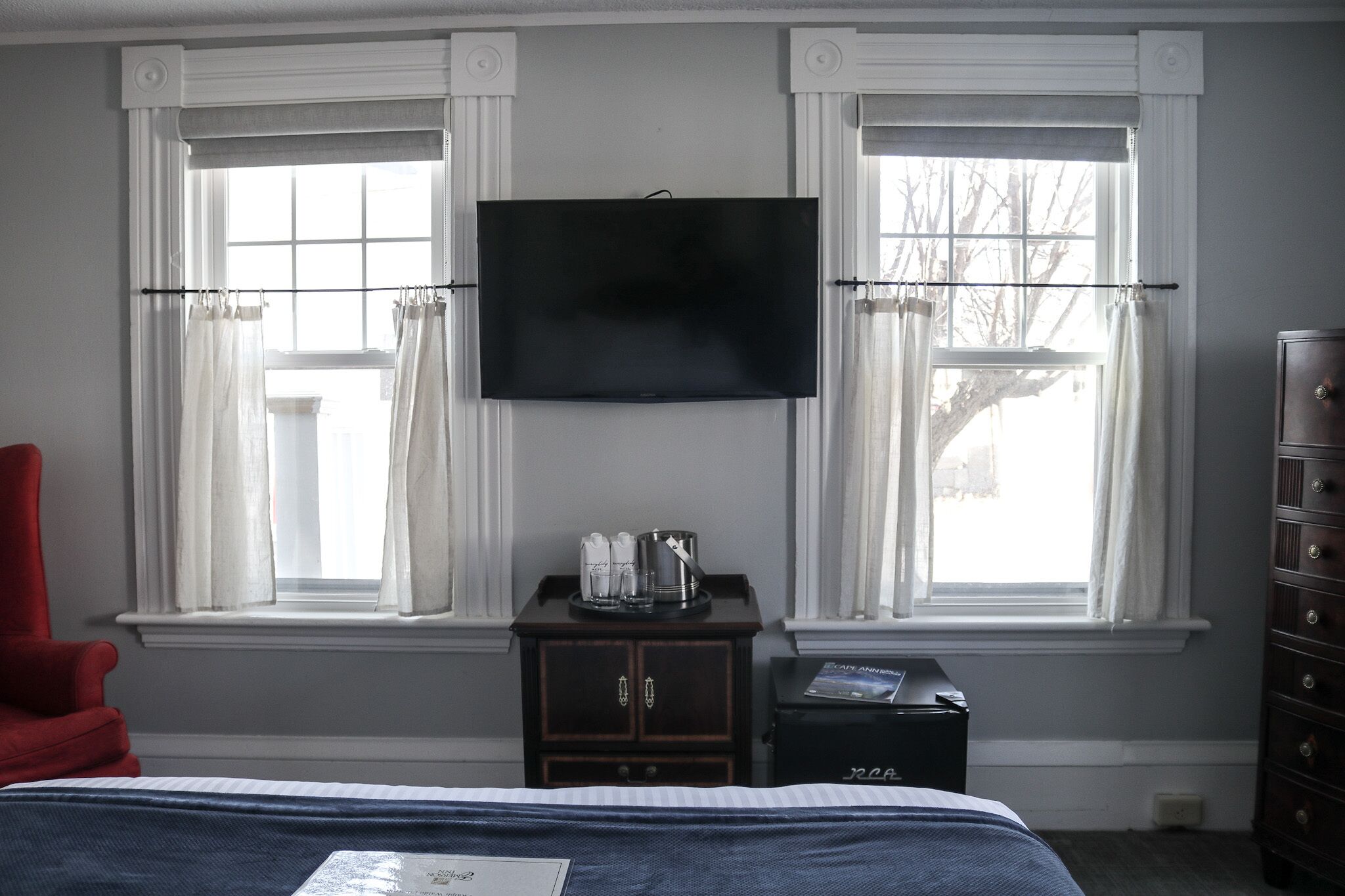 A hotel room with a bed, a TV mounted above two windows with curtains, a small cabinet with a coffee maker, and a dresser.