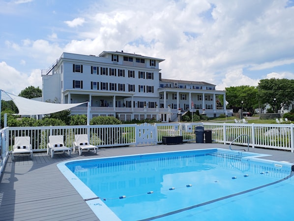 A swimming pool with a white deck, surrounded by a white fence, and a large white building with multiple balconies in the background.
