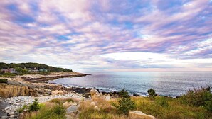 A coastal landscape with rocky terrain, a clear view of the sea, and a distant shoreline with buildings. - Emerson Inn (Rockport)