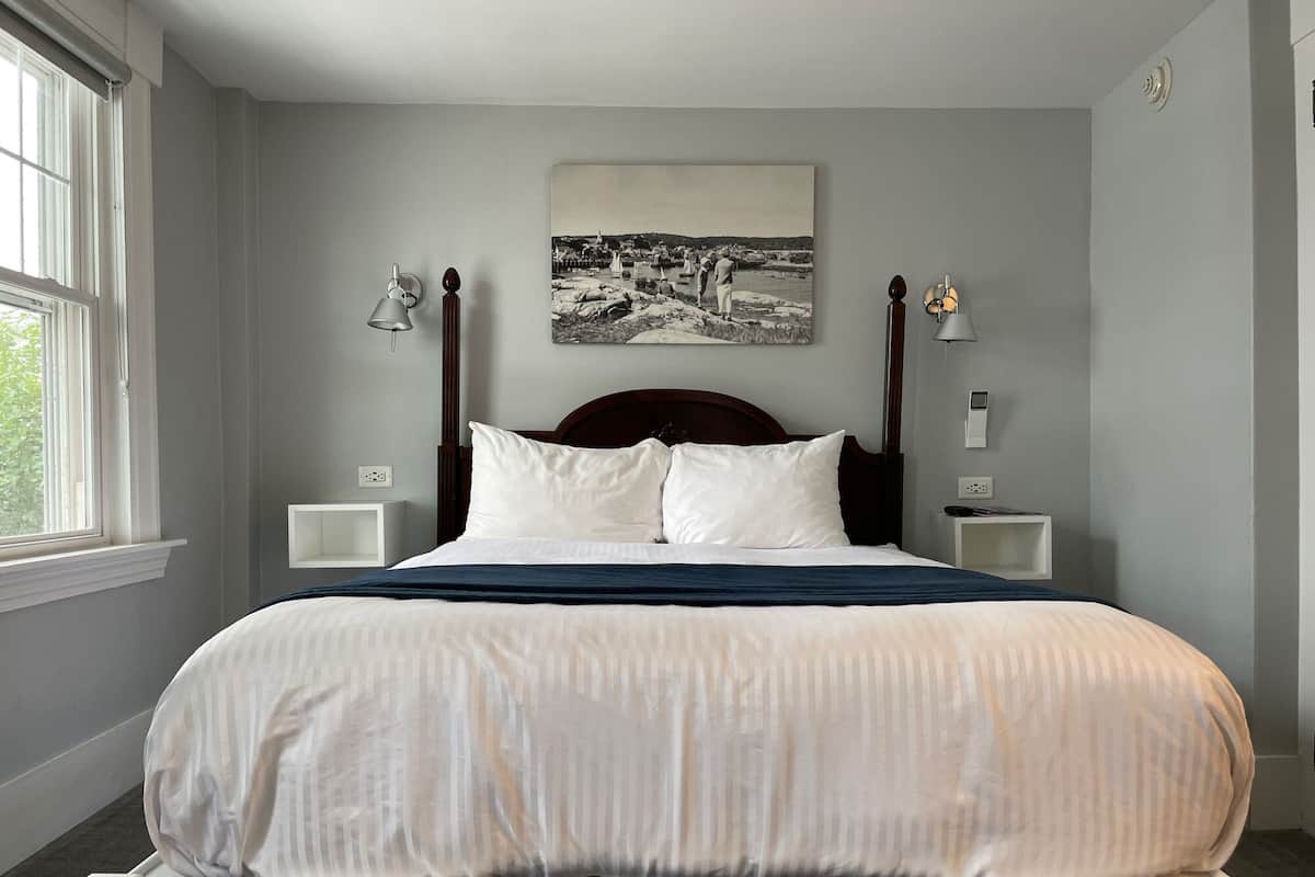 A neatly made bed with white linens and a dark blue throw blanket in a room with light gray walls. There is a black and white photograph above the bed, two bedside tables with lamps, and a window on the left side.