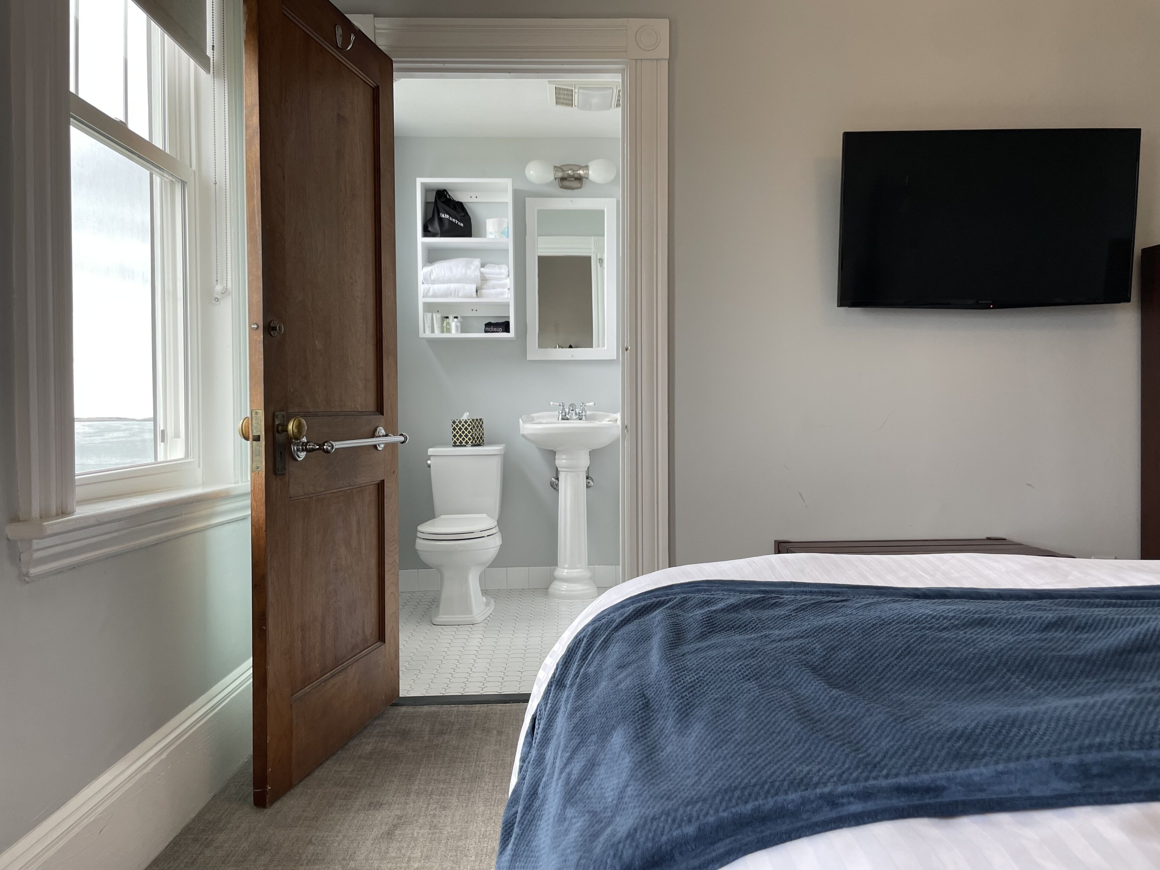 A bedroom with a bed, a wooden door, a window, a television, a bathroom with a toilet, sink, and shelves.