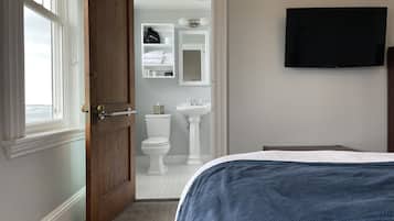 Une chambre à coucher avec un lit, une porte en bois, une fenêtre, un téléviseur, et une salle de bain comprenant des toilettes, un lavabo et des étagères.