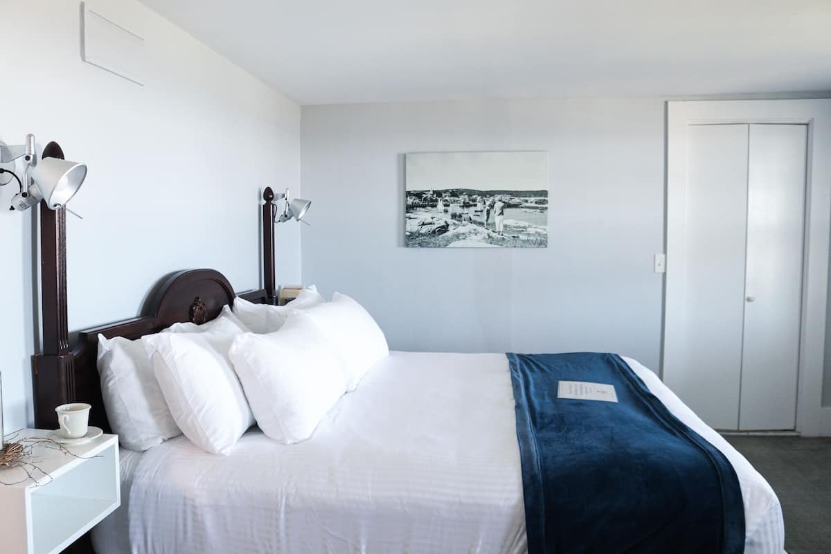 A neatly made bed with white and blue bedding, a bedside table with a cup, and a wall-mounted lamp.