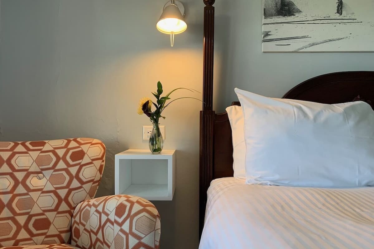 A hotel room with a bed, a nightstand with a vase of flowers, a chair, and a wall-mounted lamp.