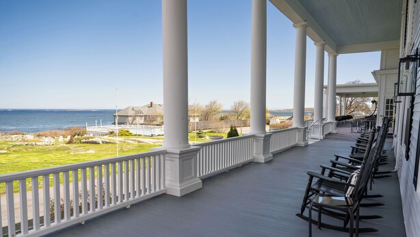 A covered porch with white columns, a row of black rocking chairs, and a view of the ocean. - Emerson Inn (Rockport)