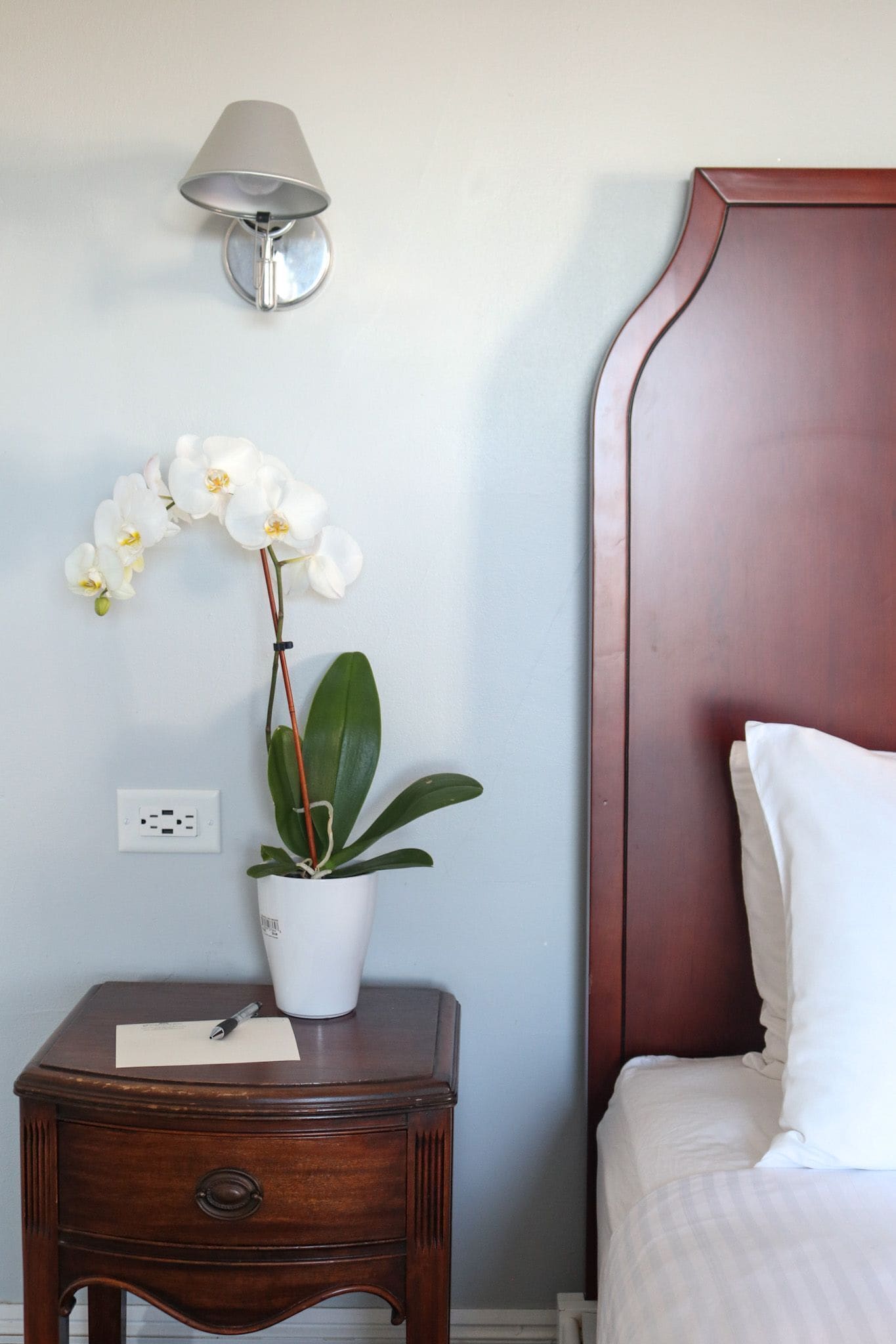 A neatly made bed with white bedding, a wooden nightstand with a white vase and an orchid, and a wall-mounted lamp.