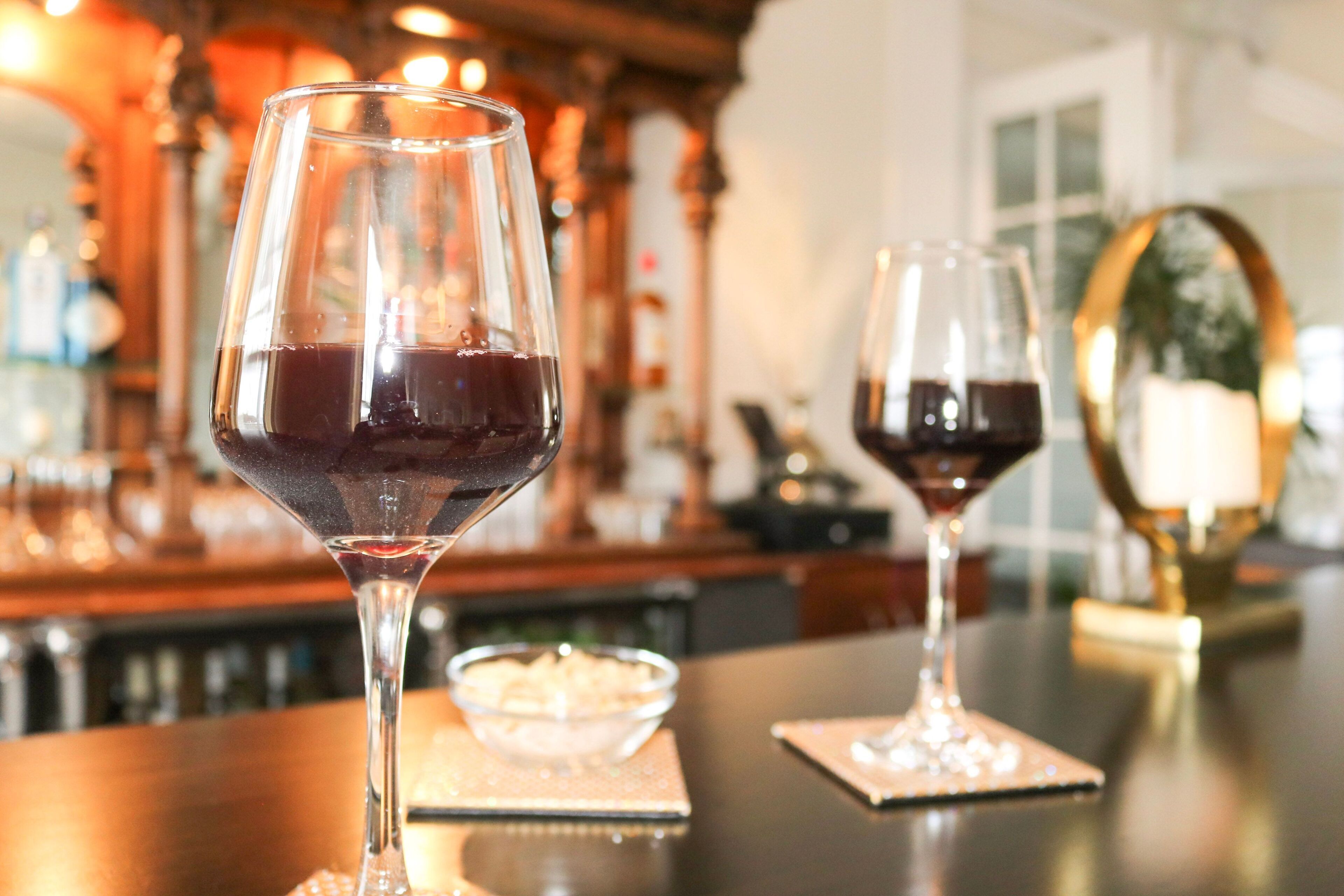 Two wine glasses on a bar counter with a candle and a bowl of snacks in the background.