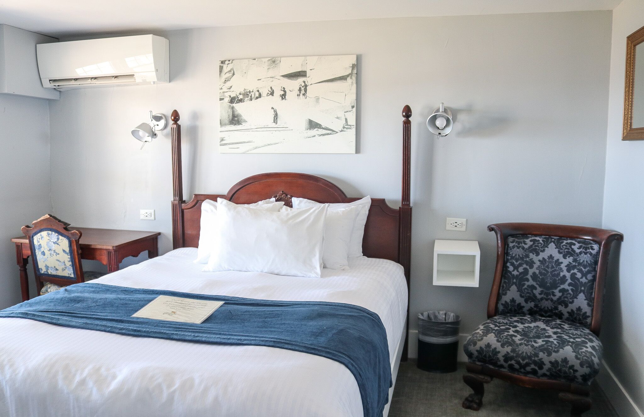 A hotel room with a wooden bed, a blue throw blanket, a framed artwork on the wall, a chair with a patterned cushion, and a trash bin.