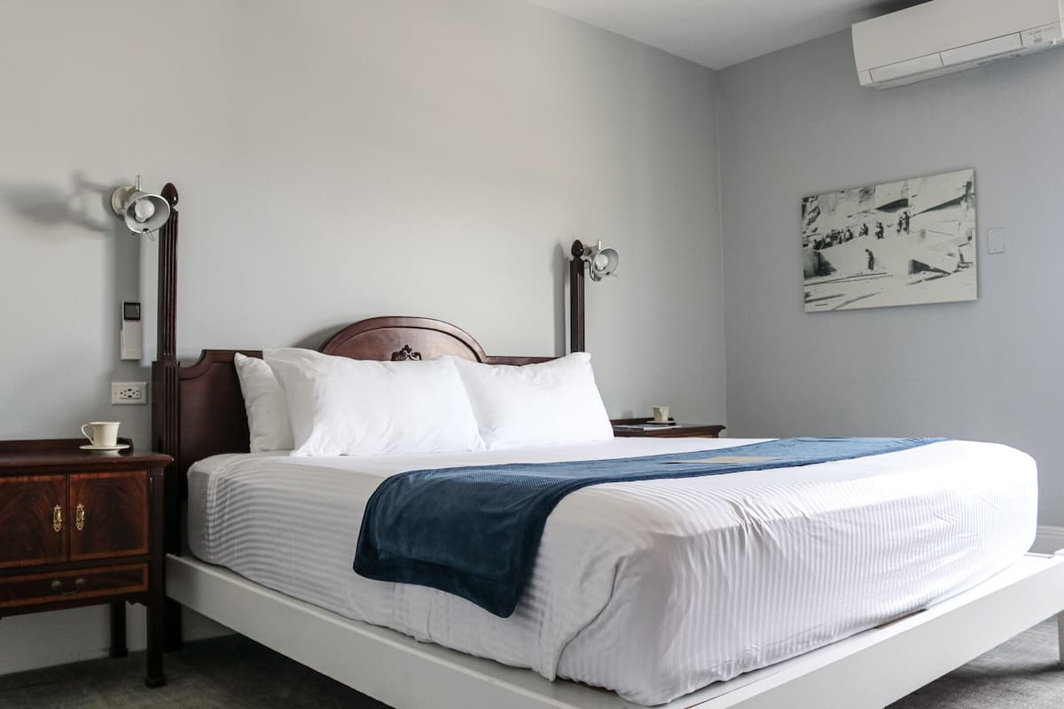 A single bed with a wooden headboard, white bedding, and a blue throw blanket. There is a nightstand with a cup and a small table lamp. A wall-mounted air conditioner and a framed abstract painting are visible.