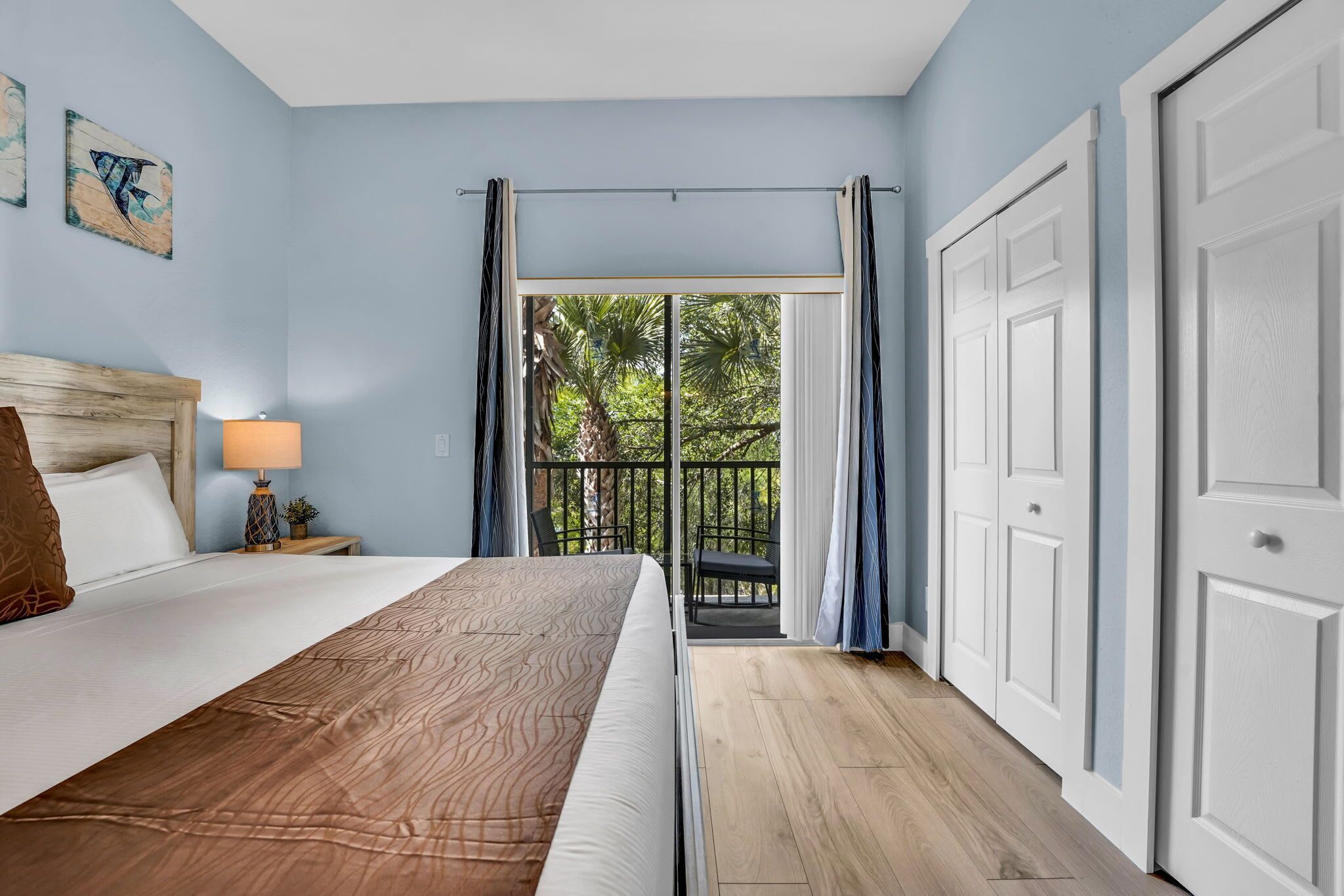 A bedroom with a bed, a nightstand with a lamp, a view of the outdoors through a balcony door, and a wooden floor.
