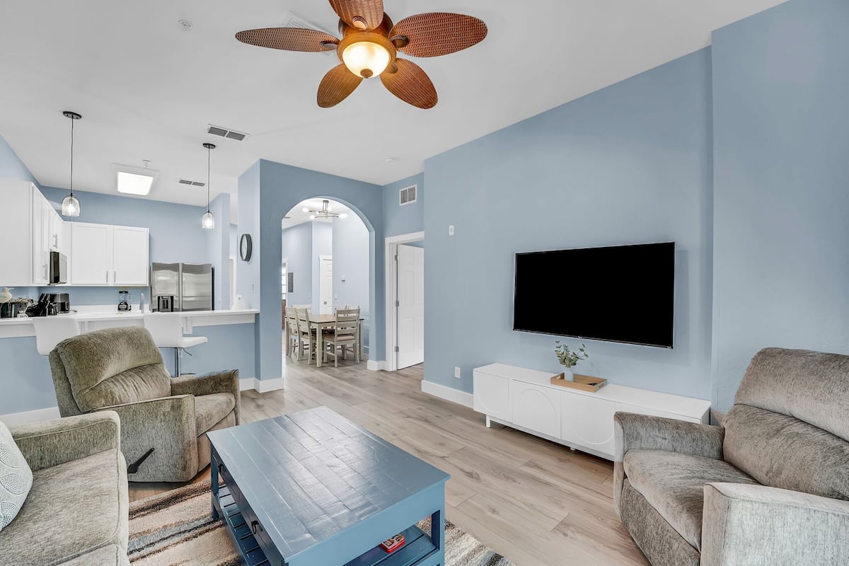 A modern living room with a flat-screen TV, a ceiling fan, and a dining area with a kitchen in the background.