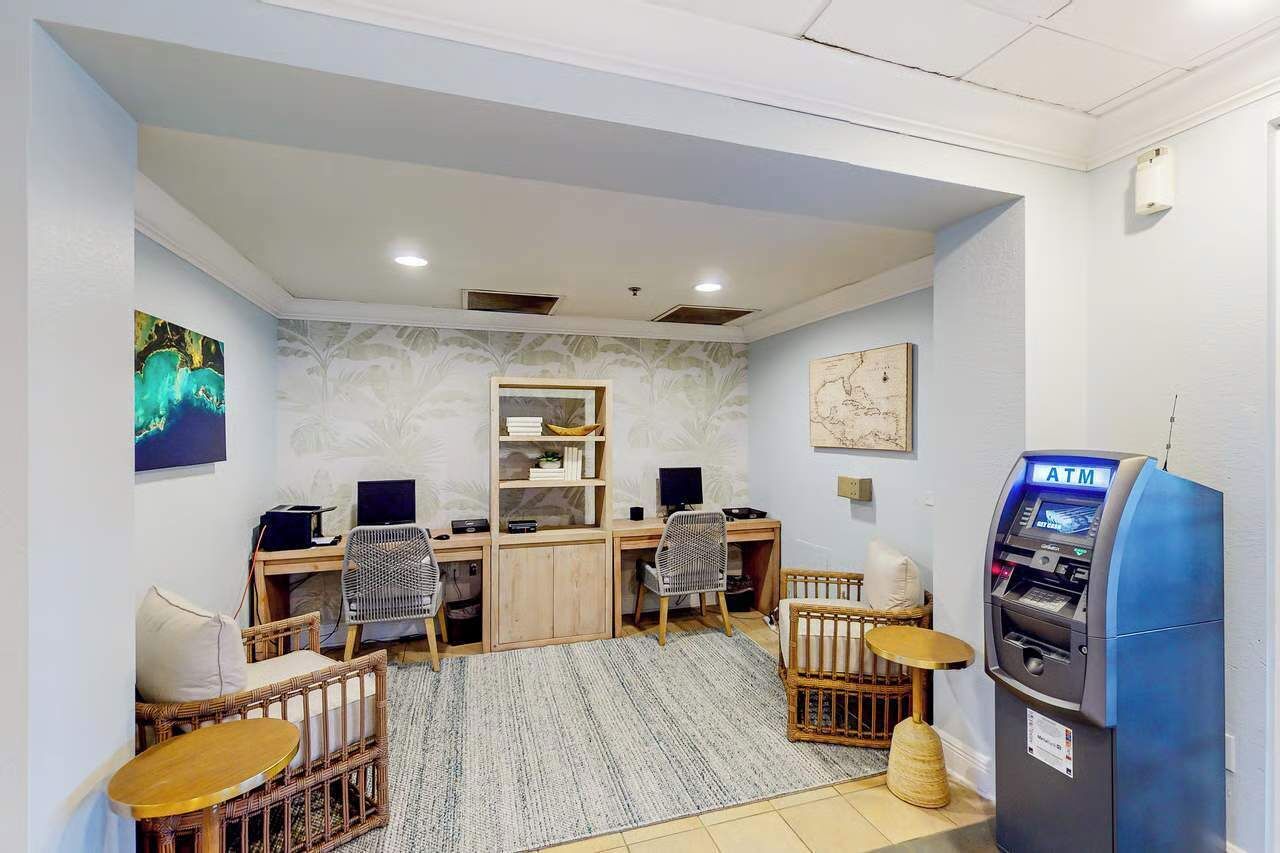 Une pièce comprenant un bureau, deux chaises, une petite table, un berceau et un distributeur automatique de billets.