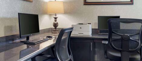 Oficina de hotel con dos monitores, una impresora, una lámpara de escritorio y una fotografía histórica enmarcada en la pared.