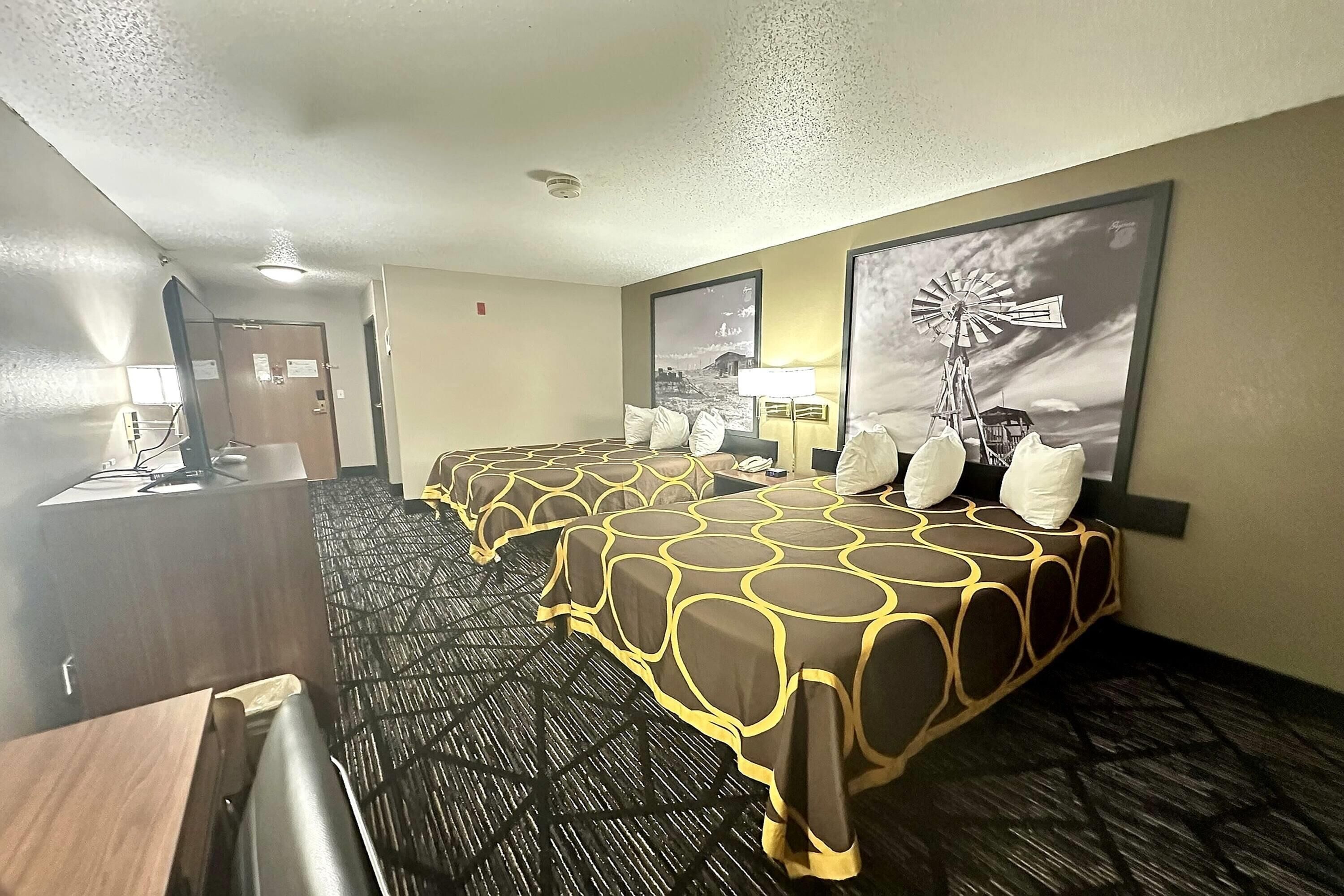 A hotel room with a long bed, a desk, and a chair. There are two wall-mounted lamps and a large window with a windmill painting.