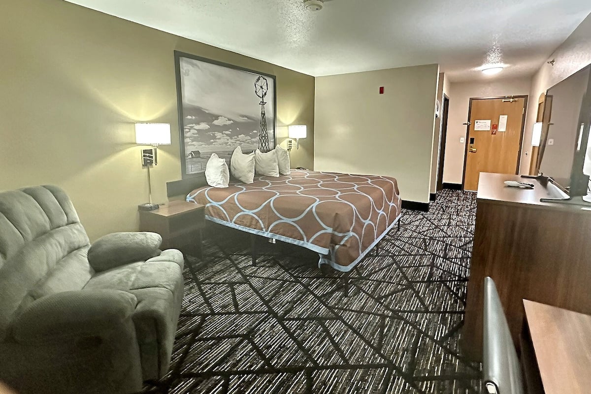 Habitación de hotel con una cama grande, una silla, un escritorio con computadora y un cuadro en la pared.