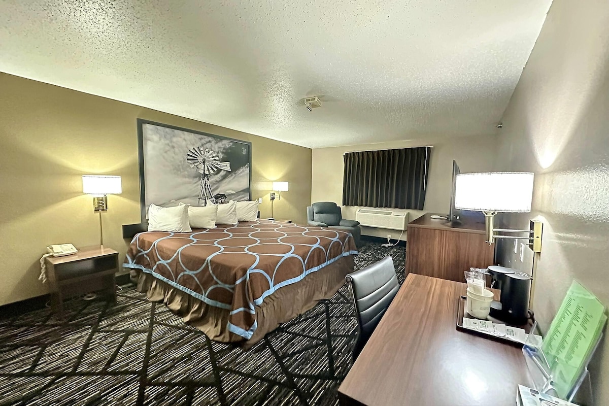 Une chambre d’hôtel avec un grand lit, un bureau, une chaise, une lampe, une horloge et une fenêtre avec des rideaux.