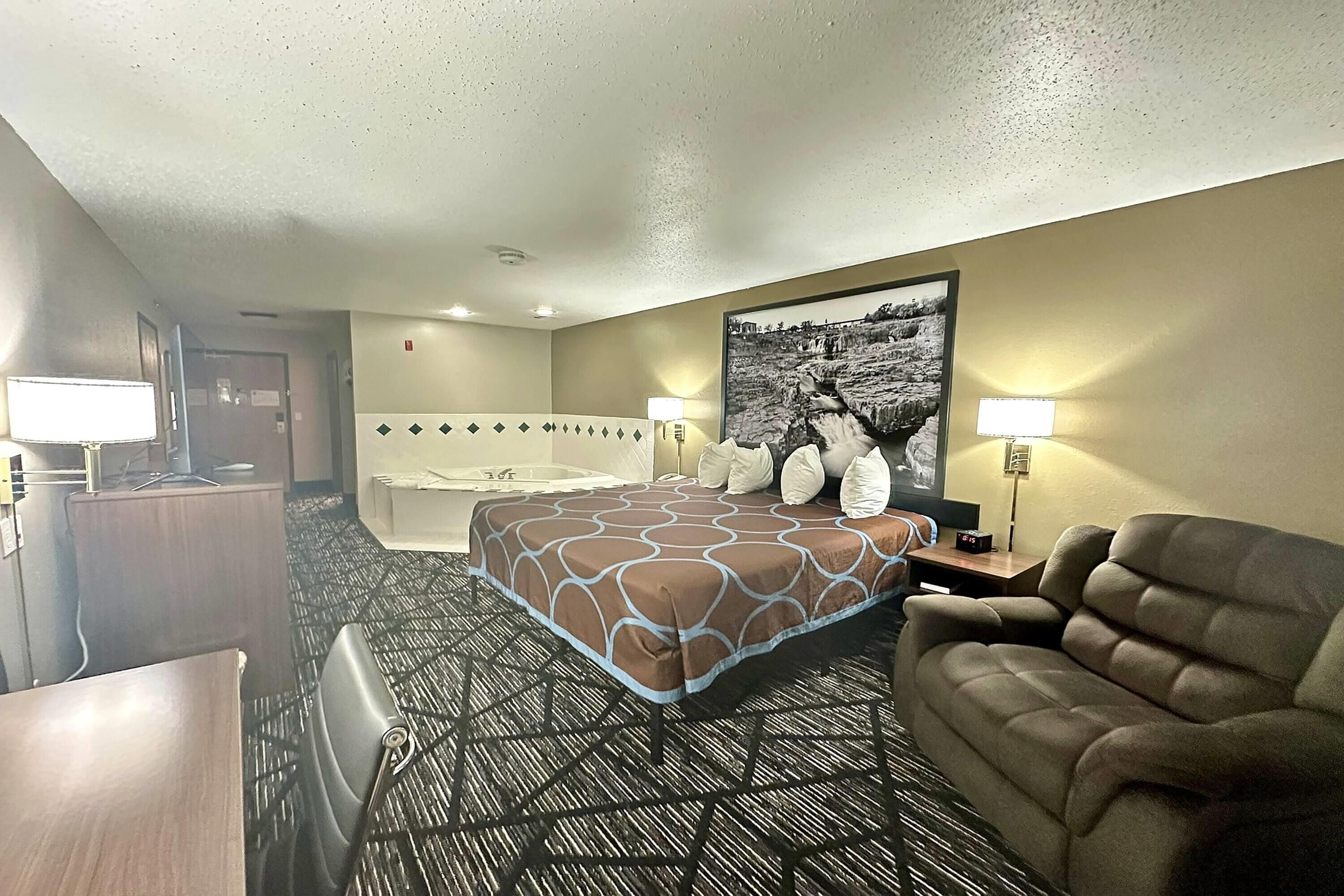 A hotel room with a bed, a sofa, a desk, a lamp, a picture on the wall, and a bathroom with a bathtub and shower.