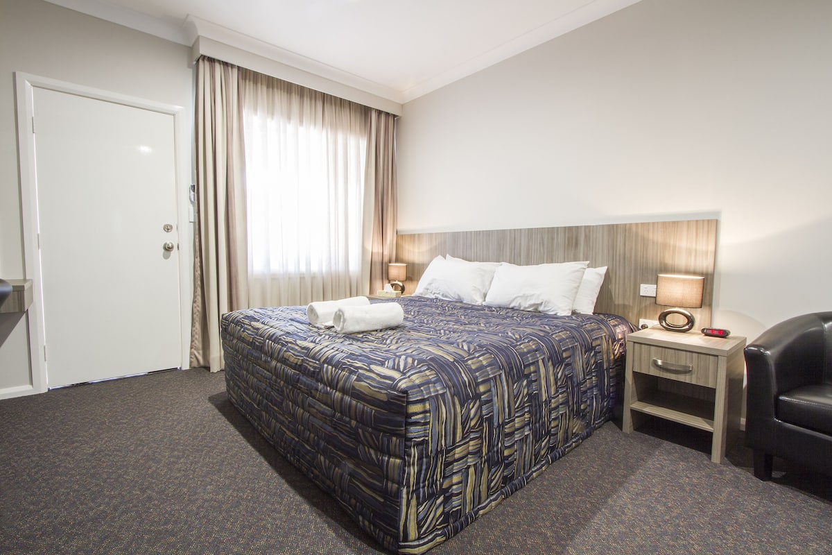 A hotel room with a large bed, a bedside table with a lamp, a chair, and a door.