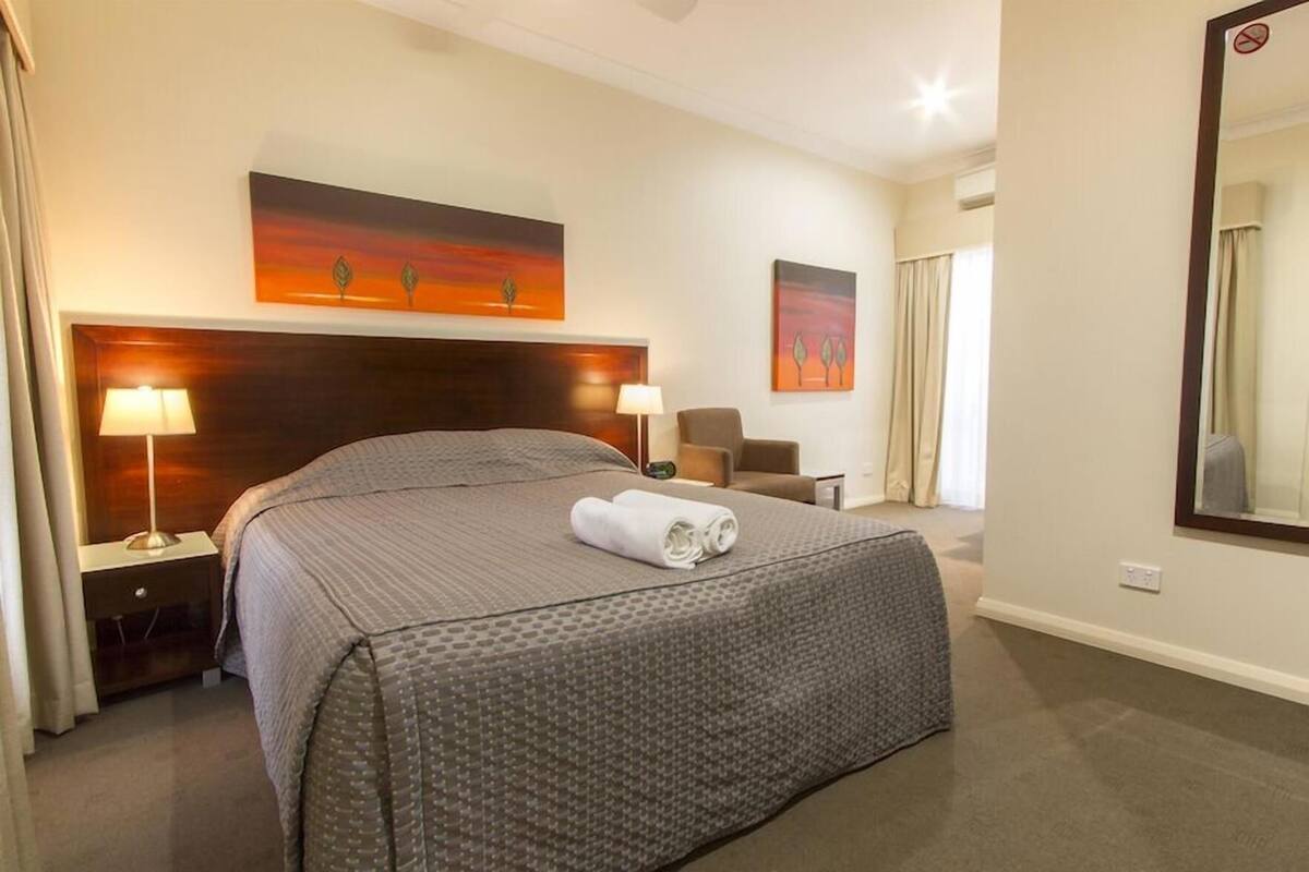 A hotel room with a large bed, two bedside lamps, a headboard, a painting above the bed, a chair, a desk, and a mirror on the wall.