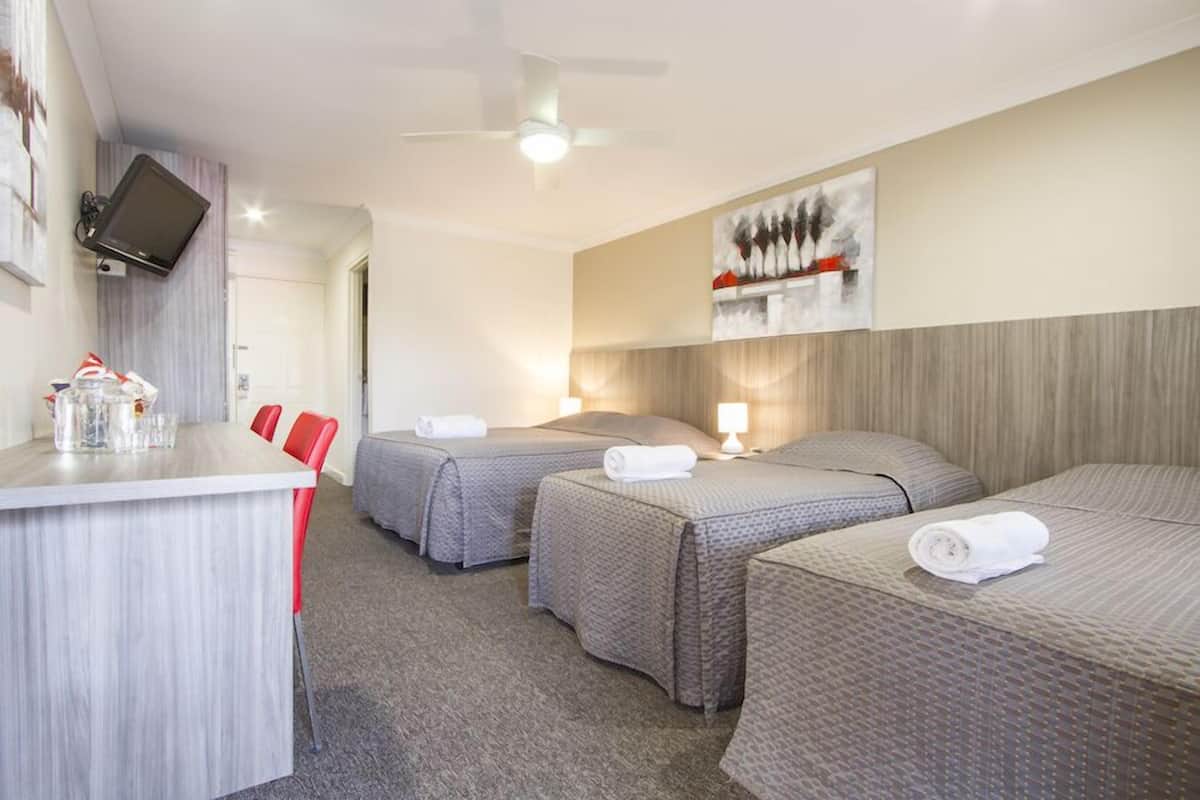 A hotel room with two single beds, a desk with a chair, a flat-screen TV mounted on the wall, and a ceiling fan.