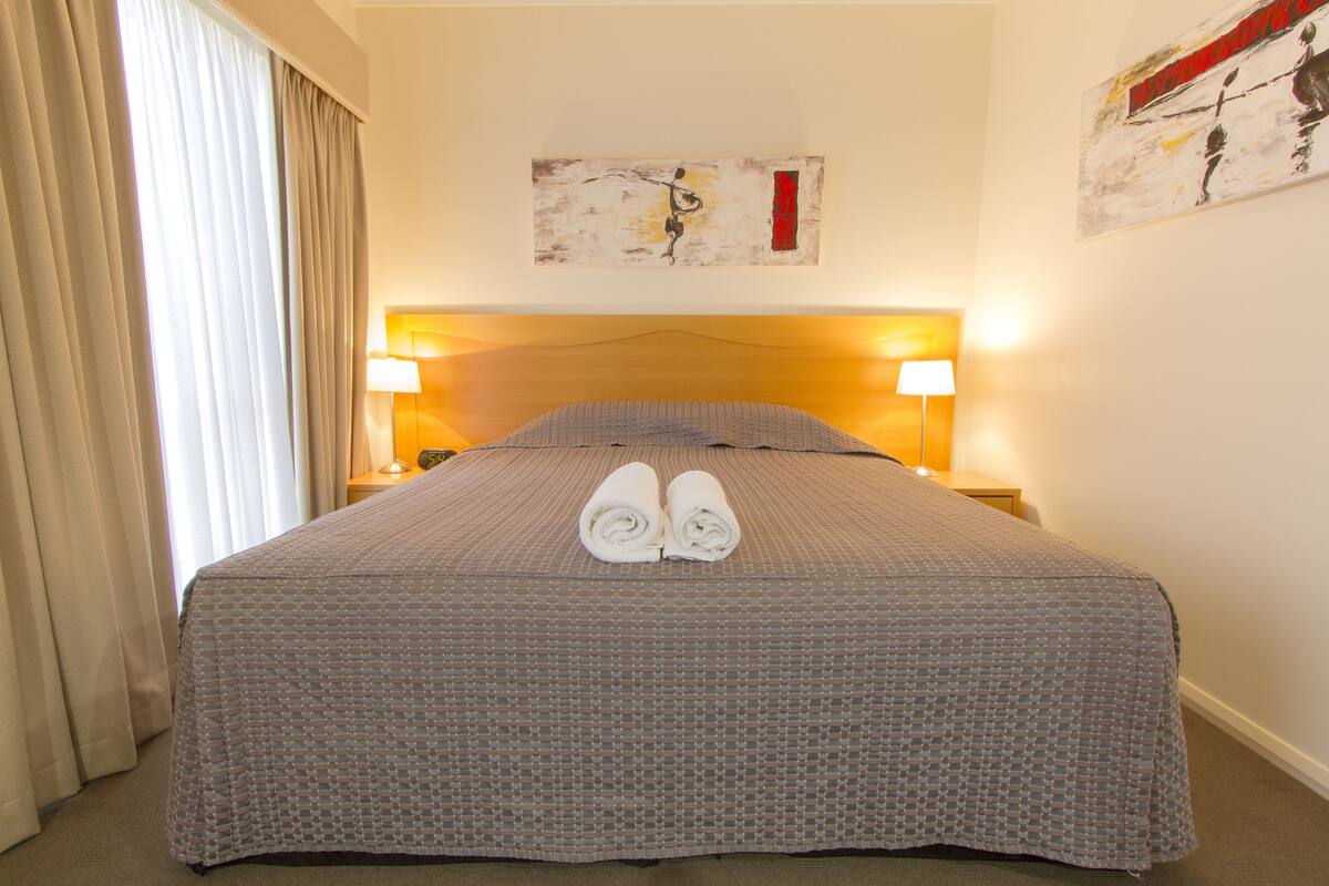 A neatly made bed with a patterned bedspread, two rolled towels on the bed, and two bedside lamps.