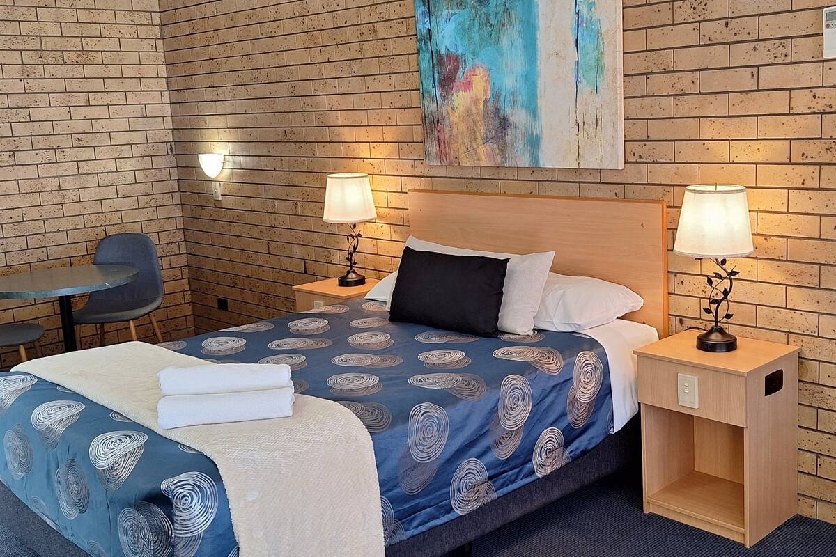 A hotel room with a brick-patterned wall, a bed with a blue patterned bedspread, two bedside tables with lamps, a chair, and a painting on the wall.