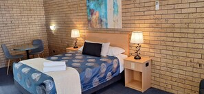 A hotel room with a brick-patterned wall, a bed with a blue patterned bedspread, two bedside tables with lamps, a chair, and a painting on the wall.
