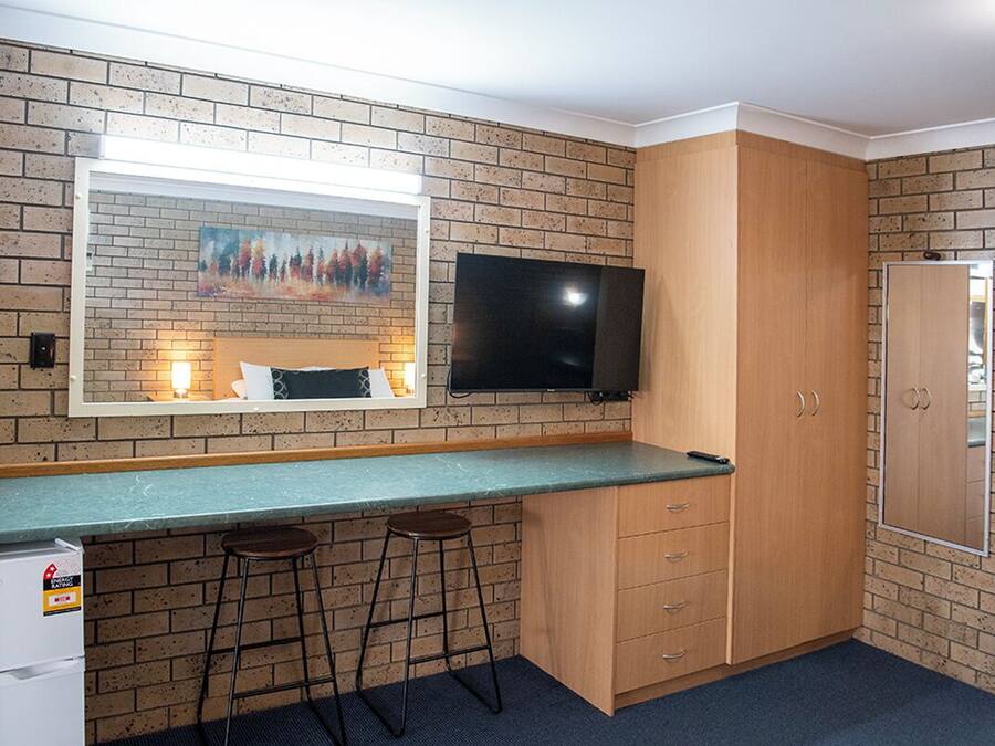 A compact kitchen with a brick backsplash, a flat-screen TV, wooden cabinets, and a bar stool.