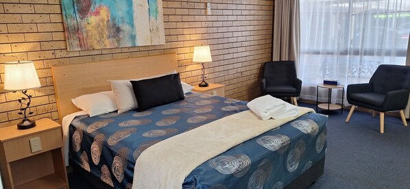 Una habitación de hotel con una pared de ladrillo, una cama con colcha estampada, dos lámparas de noche, una silla, una mesita y una ventana con cortinas.