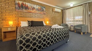 A hotel room with a brick wall, a large bed with a patterned bedspread, two bedside tables with lamps, a gray armchair, and a window with curtains. - Macintyre Motor Inn (Goondiwindi)