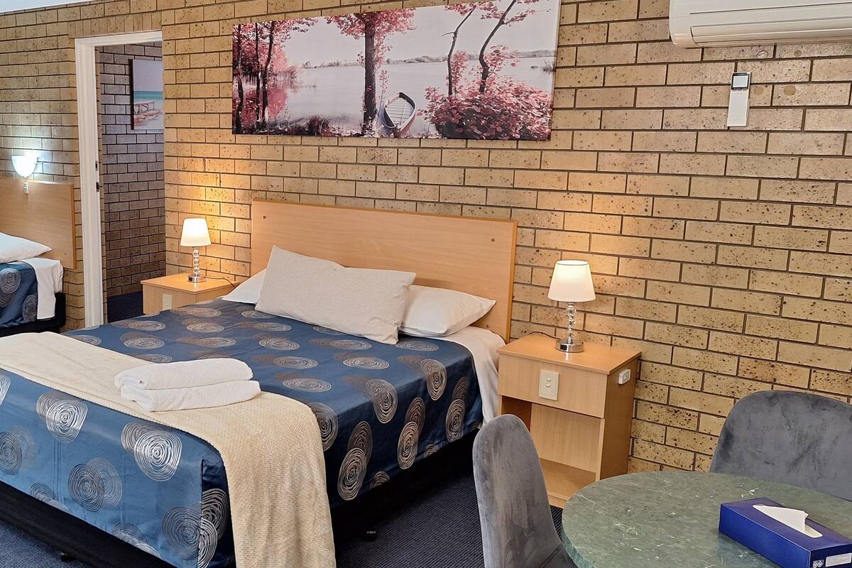A hotel room with two beds, a wooden headboard, a painting of trees, a table with a chair, and a small table with a box.