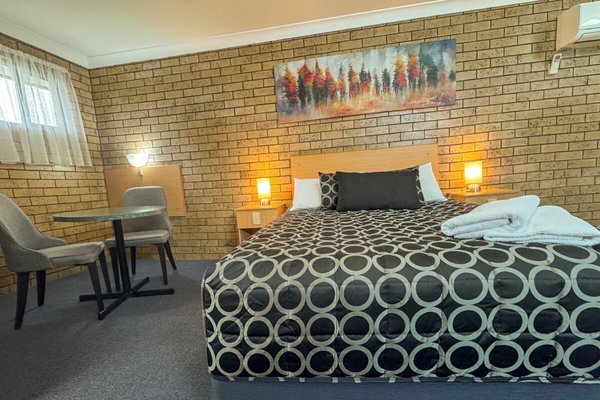 A hotel room with a brick wall, a painting of trees, a bed with a patterned bedspread, a desk with a chair, and two bedside lamps.