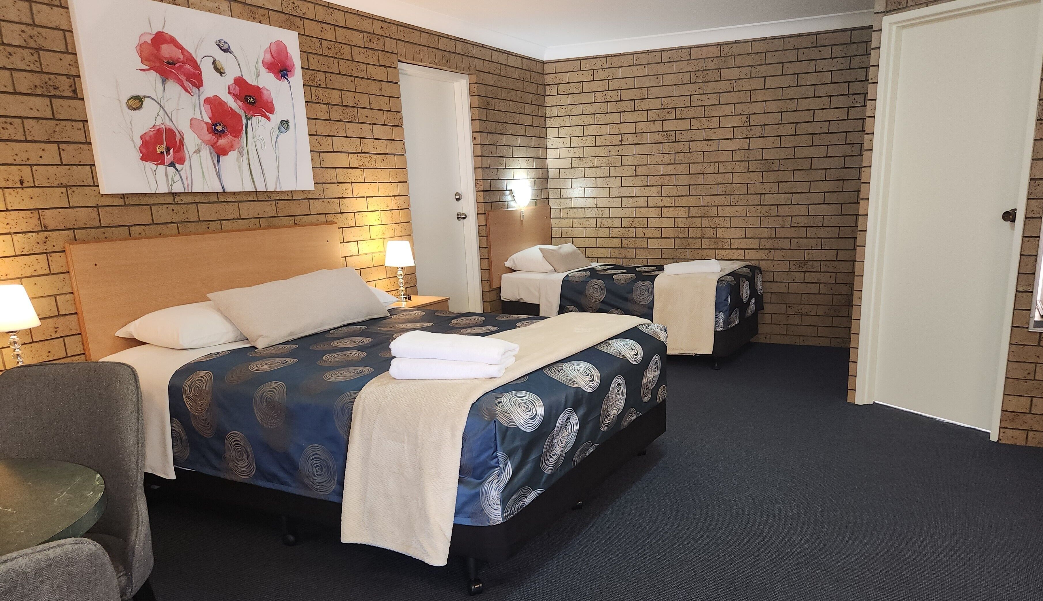 A hotel room with a brick-patterned wall, a bed with a blue and white patterned bedspread, a bedside table with a lamp, and a painting of red flowers on the wall.
