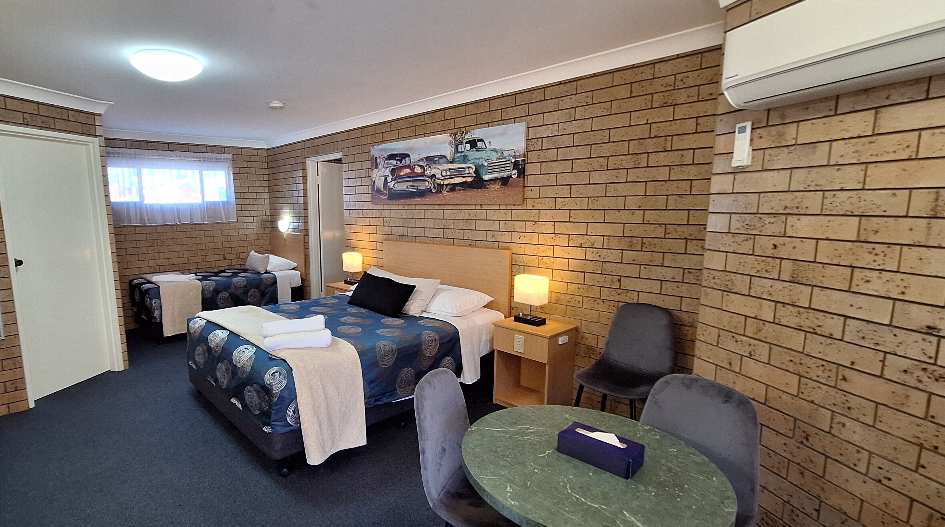 Una habitación de hotel con pared de ladrillo, una cama con colcha estampada, un escritorio con lámpara, una silla y una mesita con caja de pañuelos.