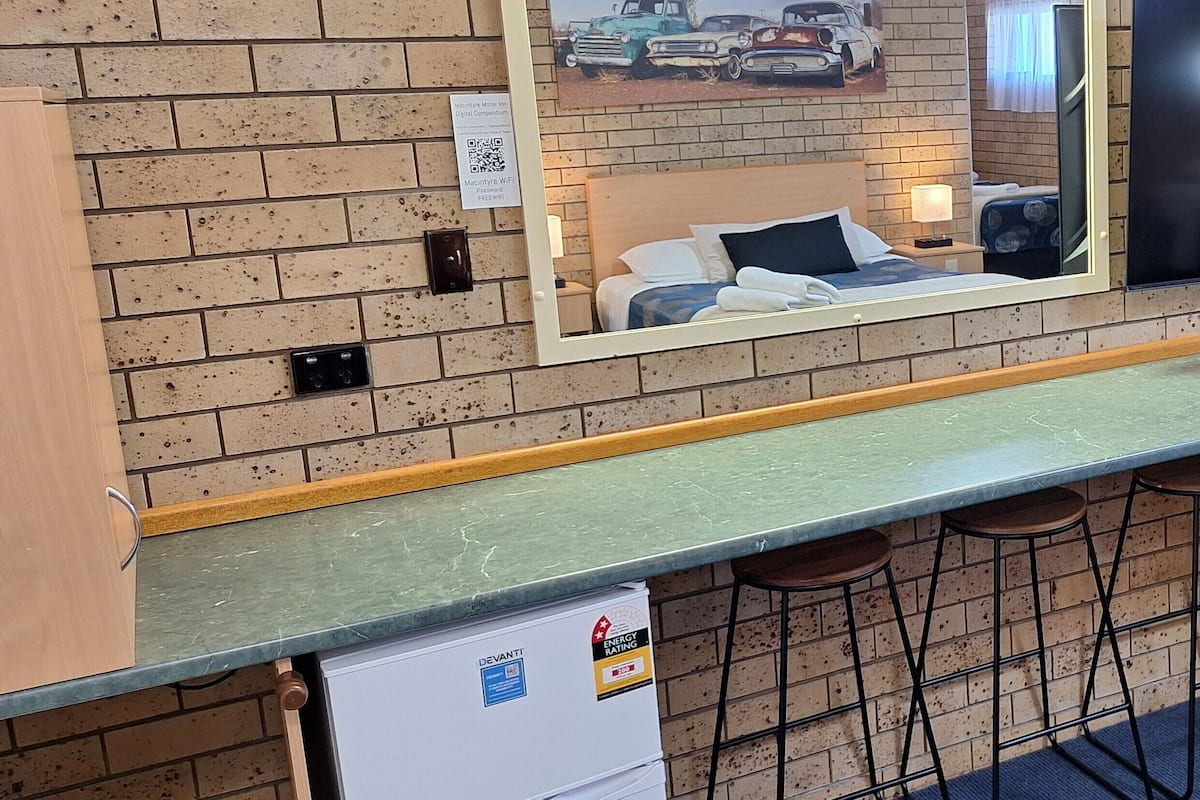 A hotel room with a brick wall, a bed with a headboard, a mirror reflecting the room, a small refrigerator, a bar area with stools, and a QR code for WiFi access.