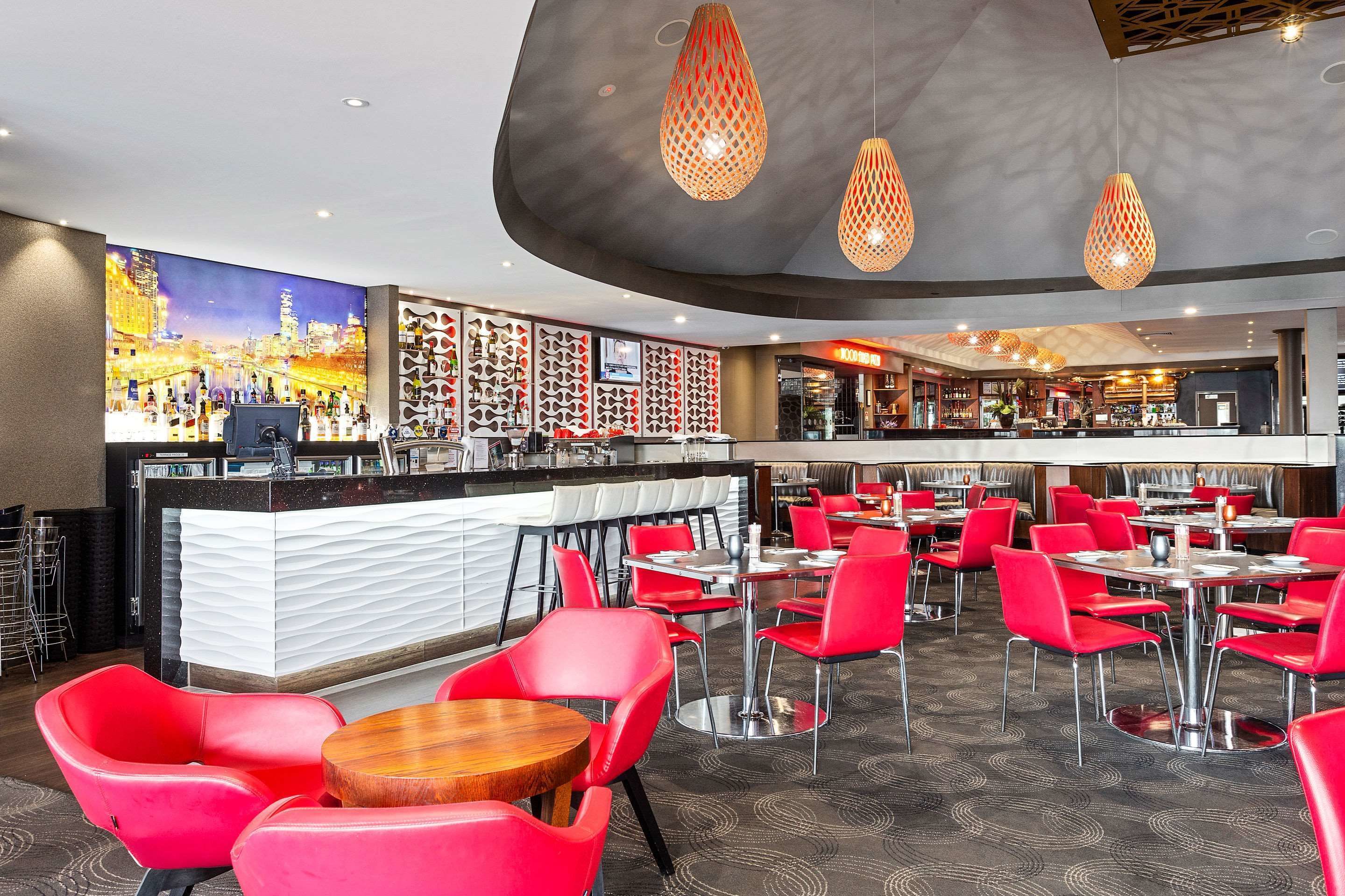A modern bar area with red chairs and tables, a counter with a white and black design, and a cityscape painting on the wall.