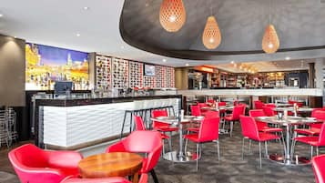 A modern bar area with red chairs and tables, a counter with a white and black design, and a cityscape painting on the wall.