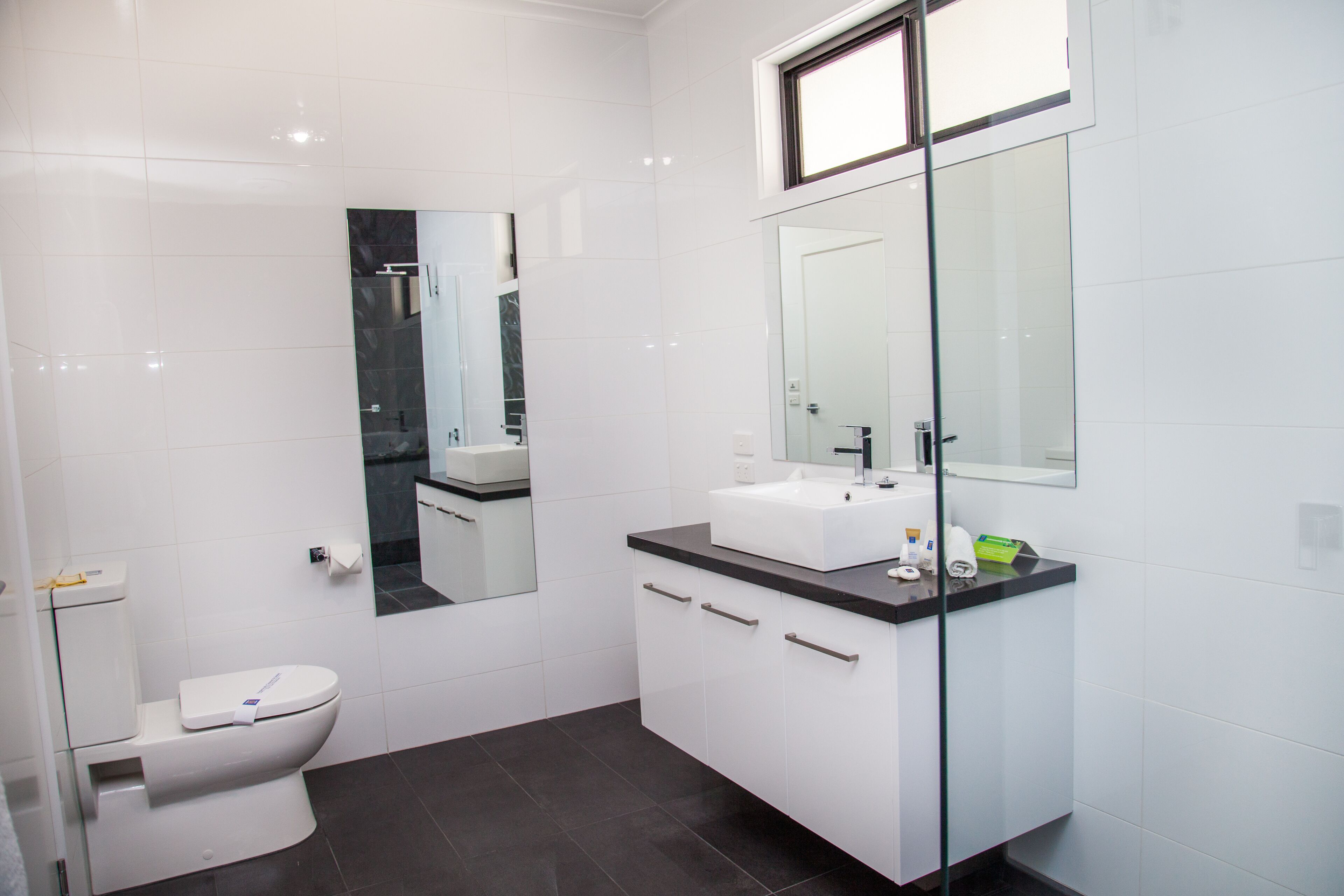 Ein modernes Badezimmer mit Toilette in Weiß, einer Duschkabine aus Glas, einem weißen Waschbecken mit schwarzer Arbeitsplatte und einem großen Spiegel über dem Waschbecken.