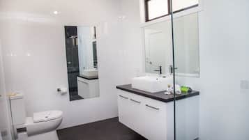Ein modernes Badezimmer mit Toilette in Weiß, einer Duschkabine aus Glas, einem weißen Waschbecken mit schwarzer Arbeitsplatte und einem großen Spiegel über dem Waschbecken.