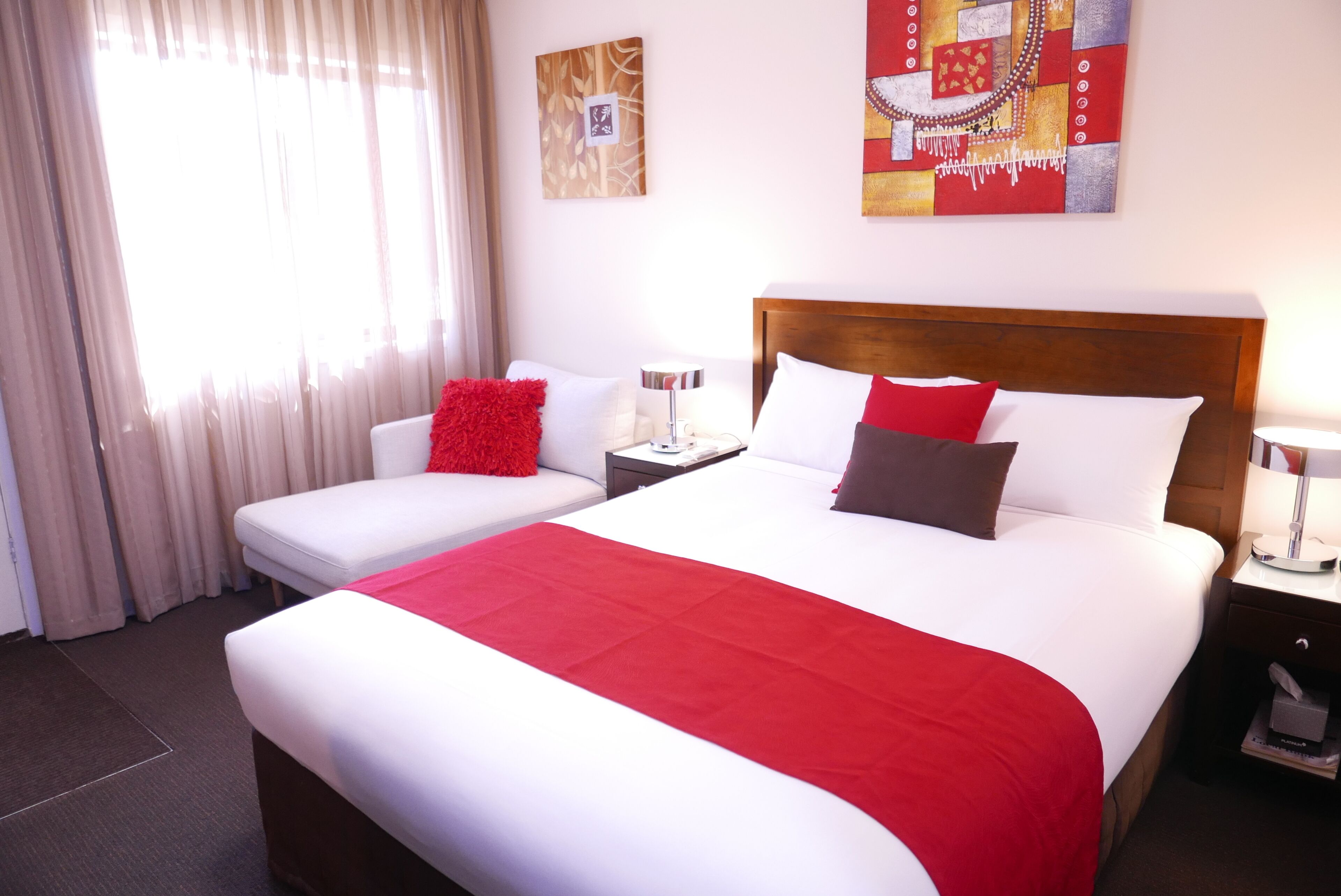 A hotel room with a large bed, a smaller bed, a nightstand with a lamp, a window with curtains, and artwork on the walls.