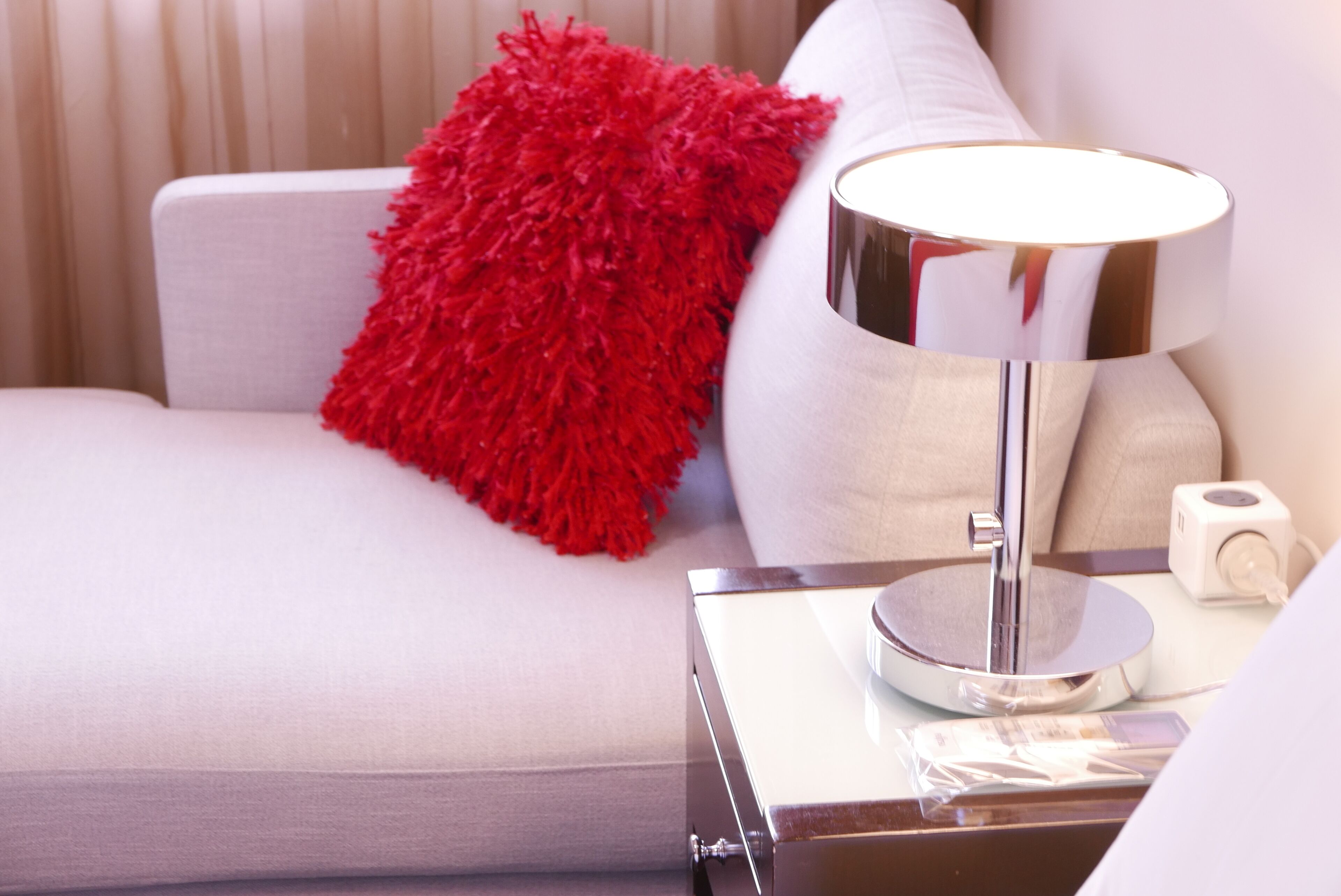 A modern bedroom with a white sofa, a red fluffy pillow, a chrome bedside table with a lamp, and a small white device on the table.