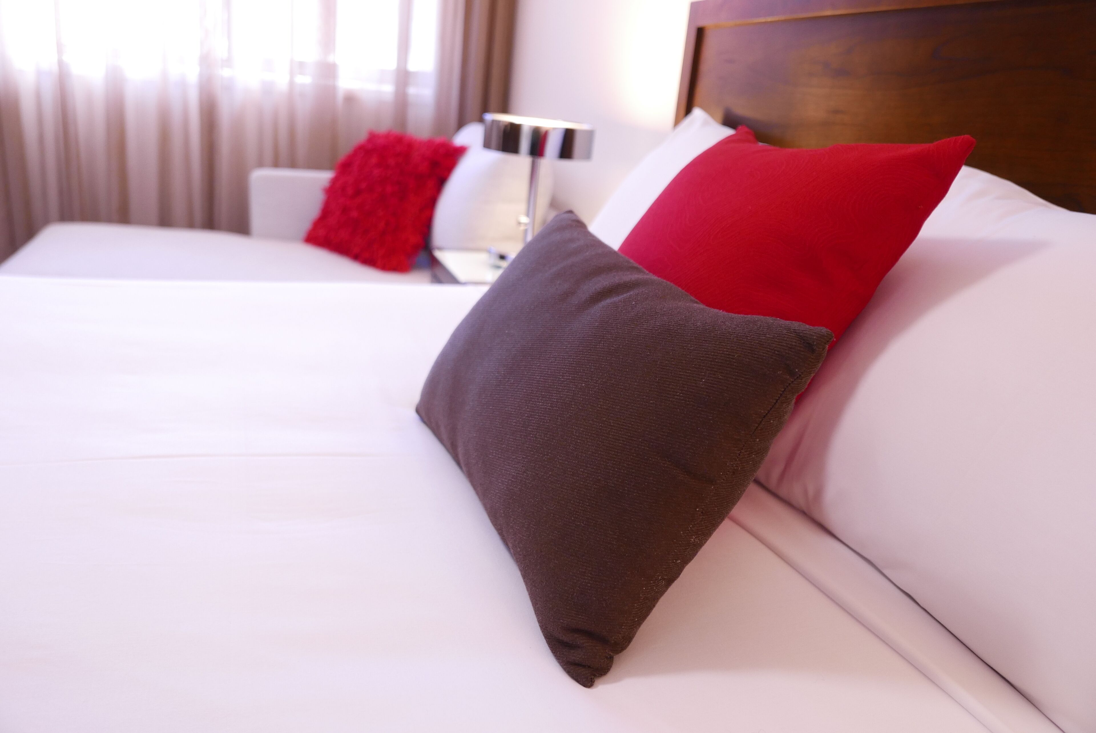 A neatly made bed with white bedding and two pillows, one red and one brown, in a hotel room with a wooden headboard and a bedside table with a lamp.